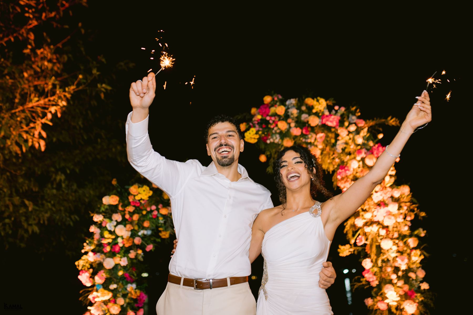 Anas and Salma holding sparklers in front of a flower arch at night